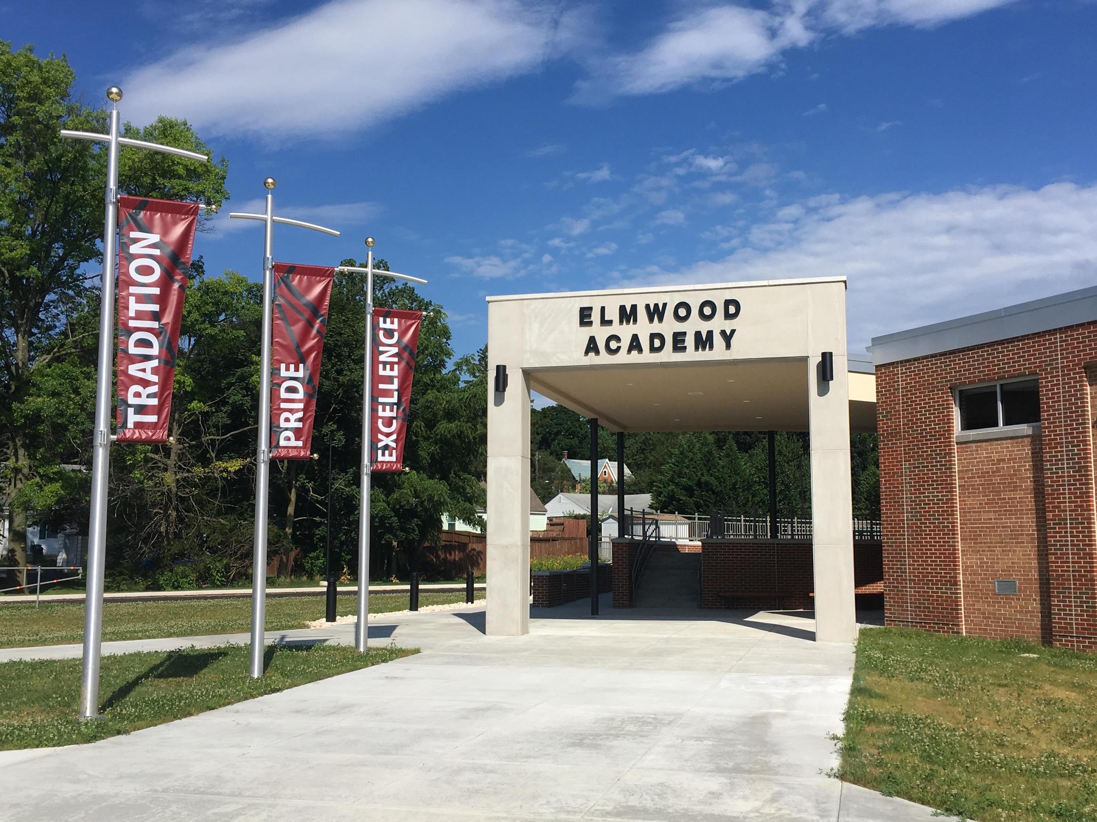 The entrance of Elmwood Academy features a covered walkway with the name 'ELMWOOD ACADEMY' prominently displayed, flanked by banners that read 'TRADITION', 'PRIDE', and 'EXCELLENCE', set against a clear blue sky.