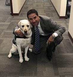 A man in a suit kneels beside a yellow Labrador retriever wearing a harness, both smiling in an office hallway with gray carpet.