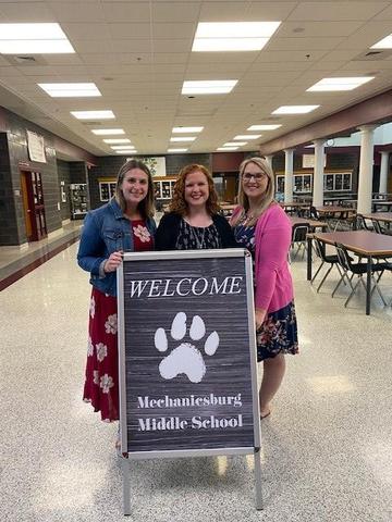 Three women stand in a school cafeteria next to a welcome sign for Mechanicsburg Middle School, with tables and chairs visible in the background.
