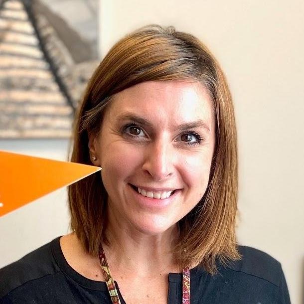 A smiling woman with shoulder-length brown hair, wearing a black top, stands in front of a neutral background, holding an orange triangular flag.