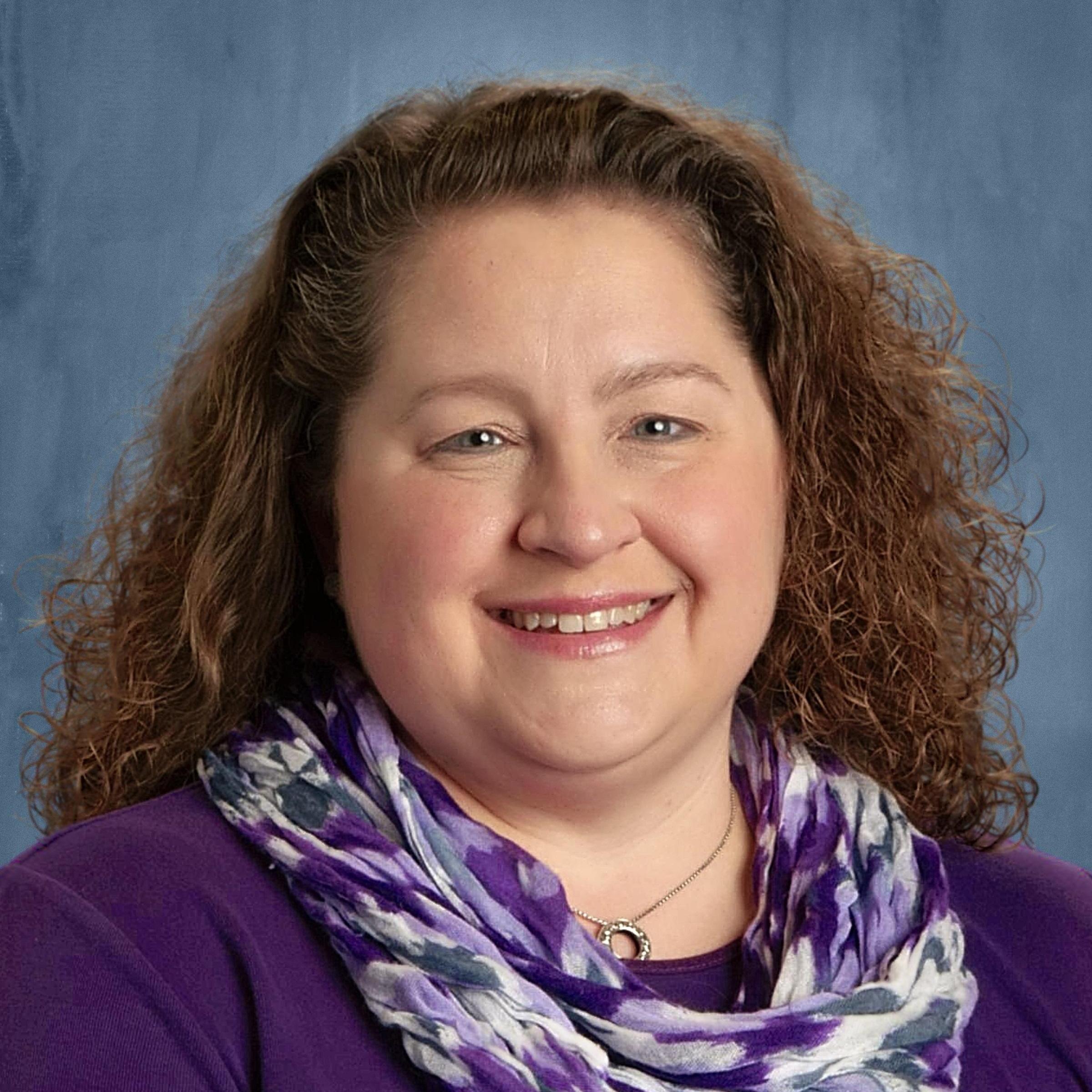 A smiling woman with curly hair, wearing a purple top and a patterned scarf, poses against a blue background.