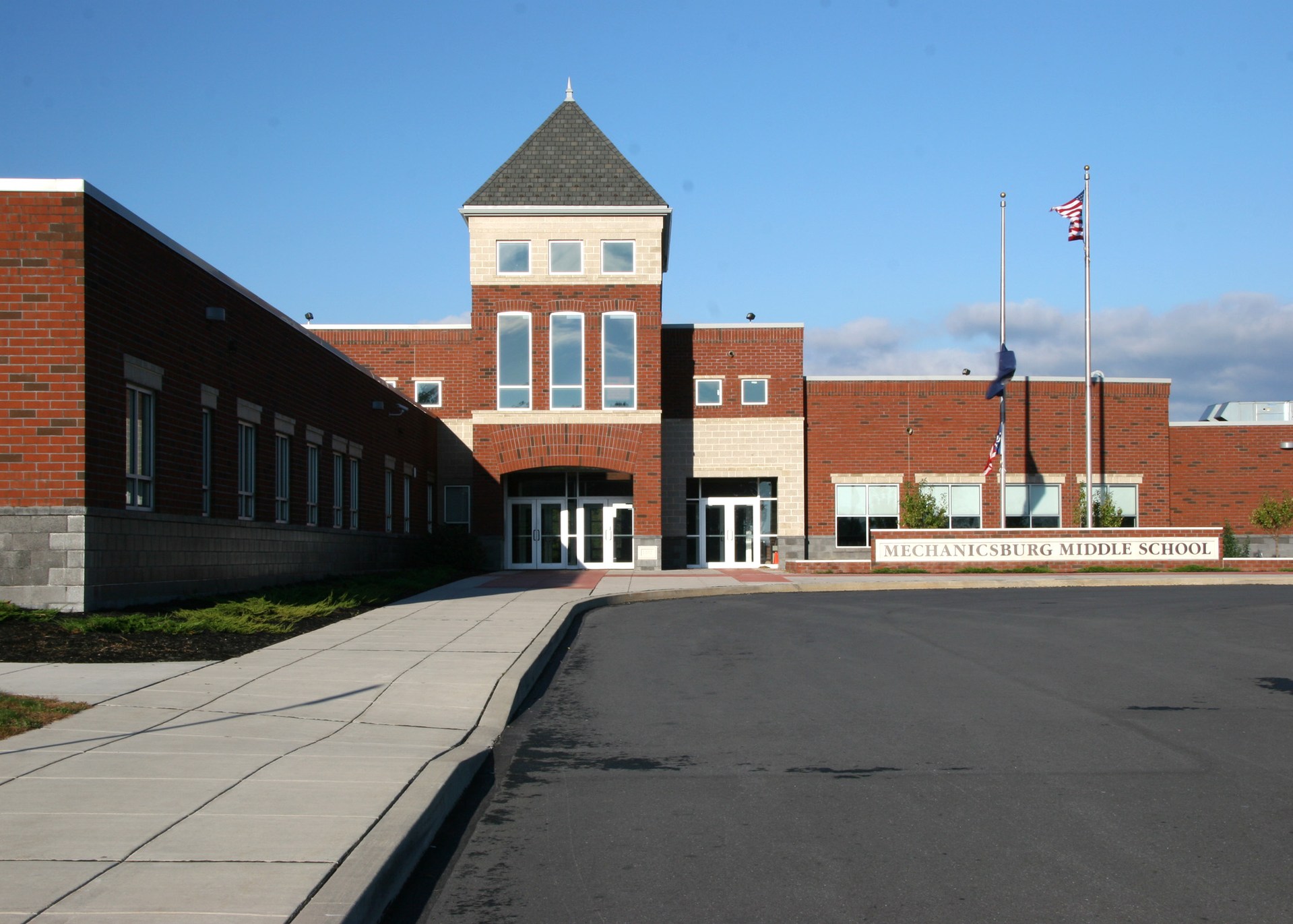 A brick school building with a central tower, flanked by large windows and a paved walkway leading to the entrance, under a clear blue sky.
