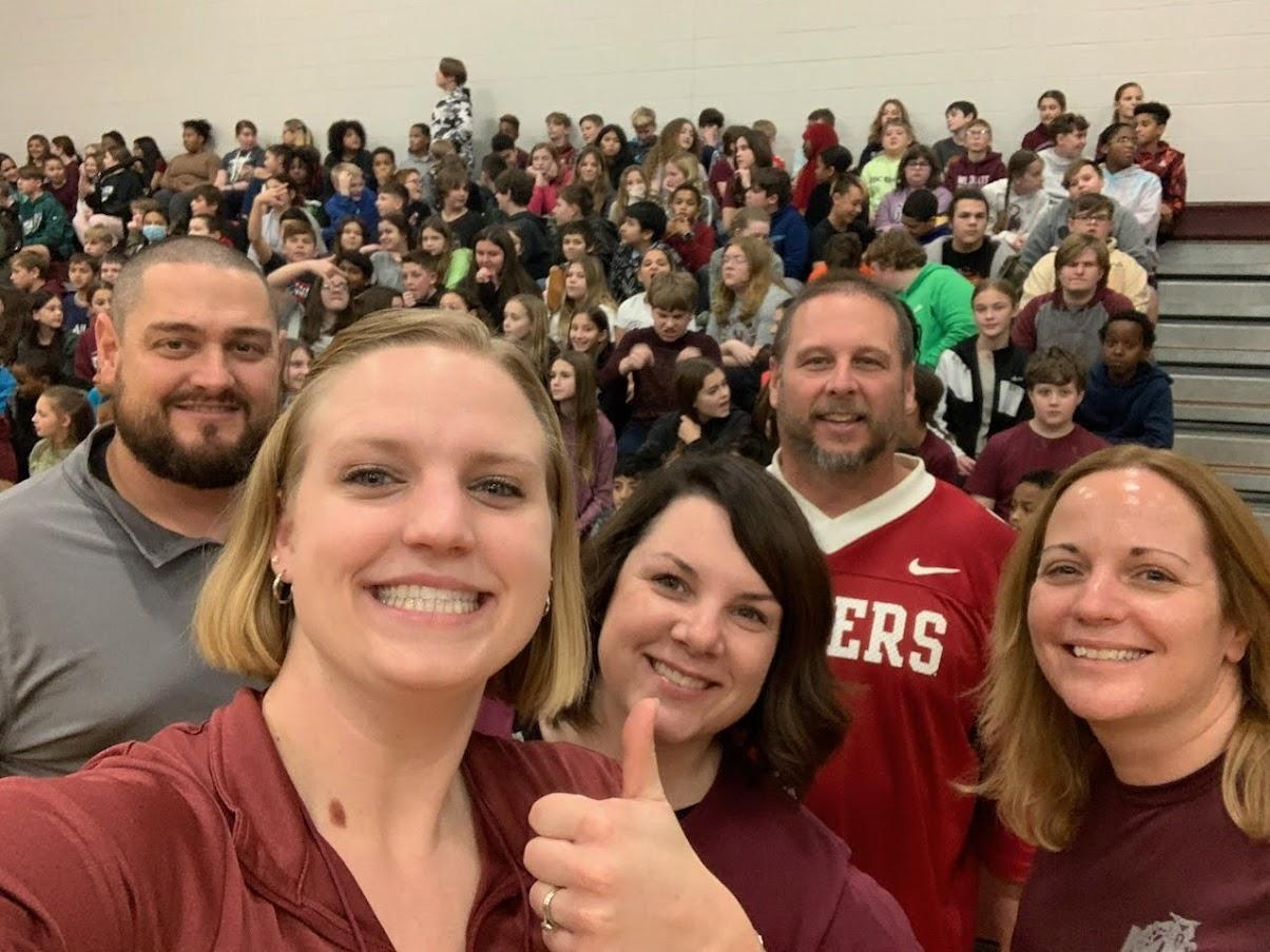 A group of five smiling individuals pose for a selfie in front of a large crowd of students seated in a gymnasium.