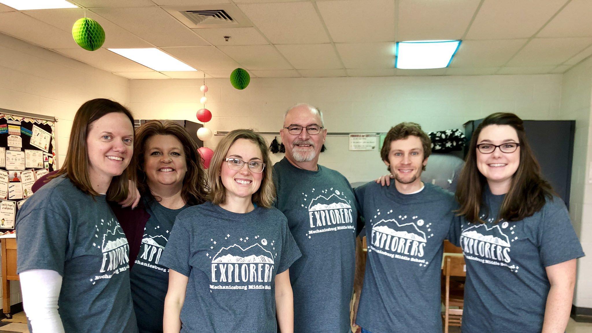A group of six individuals wearing matching gray t-shirts with a design, standing together in a classroom setting decorated with colorful hanging ornaments.