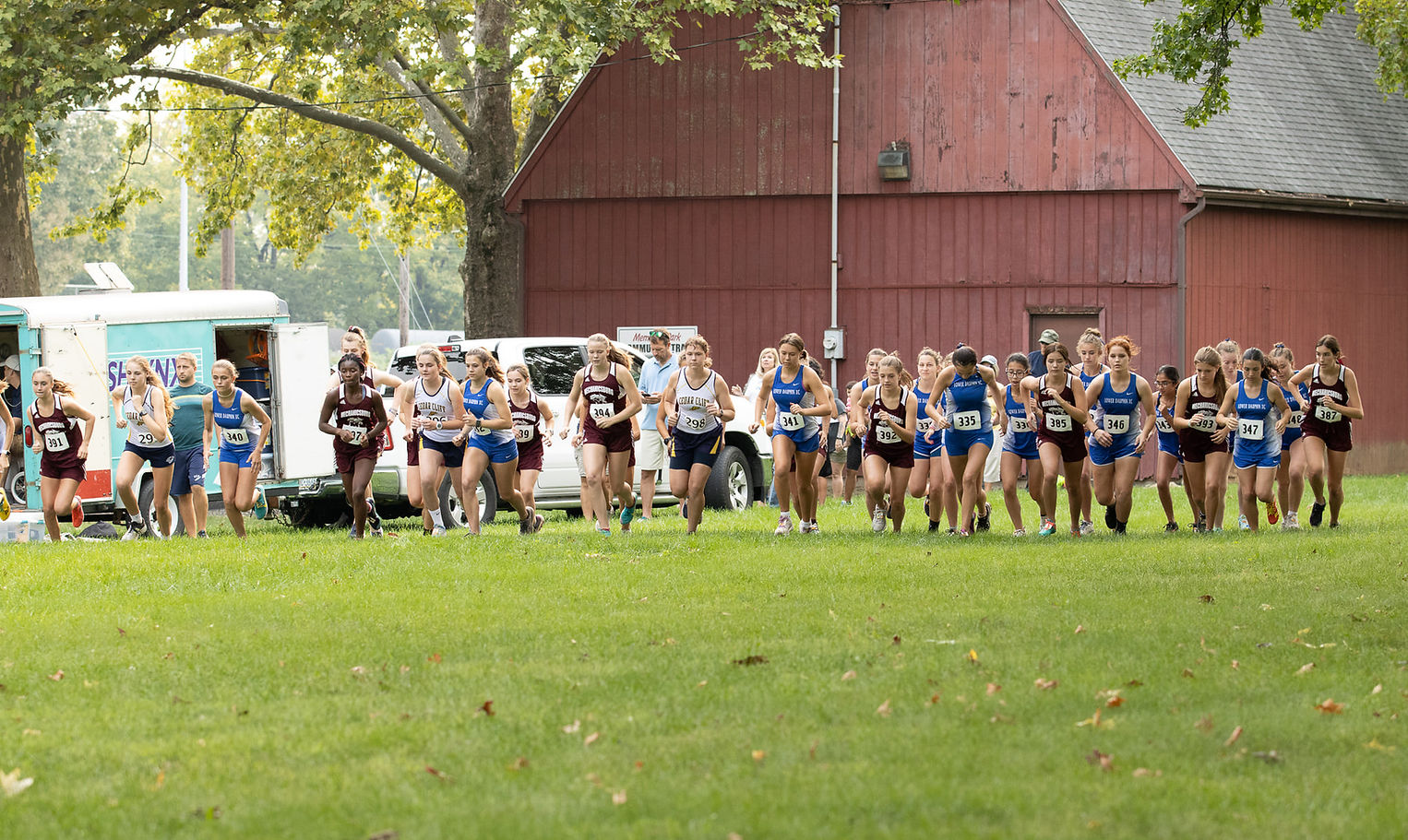 A group of female cross country runners in athletic gear are starting a race on a grassy field, with a barn and vehicles visible in the background.