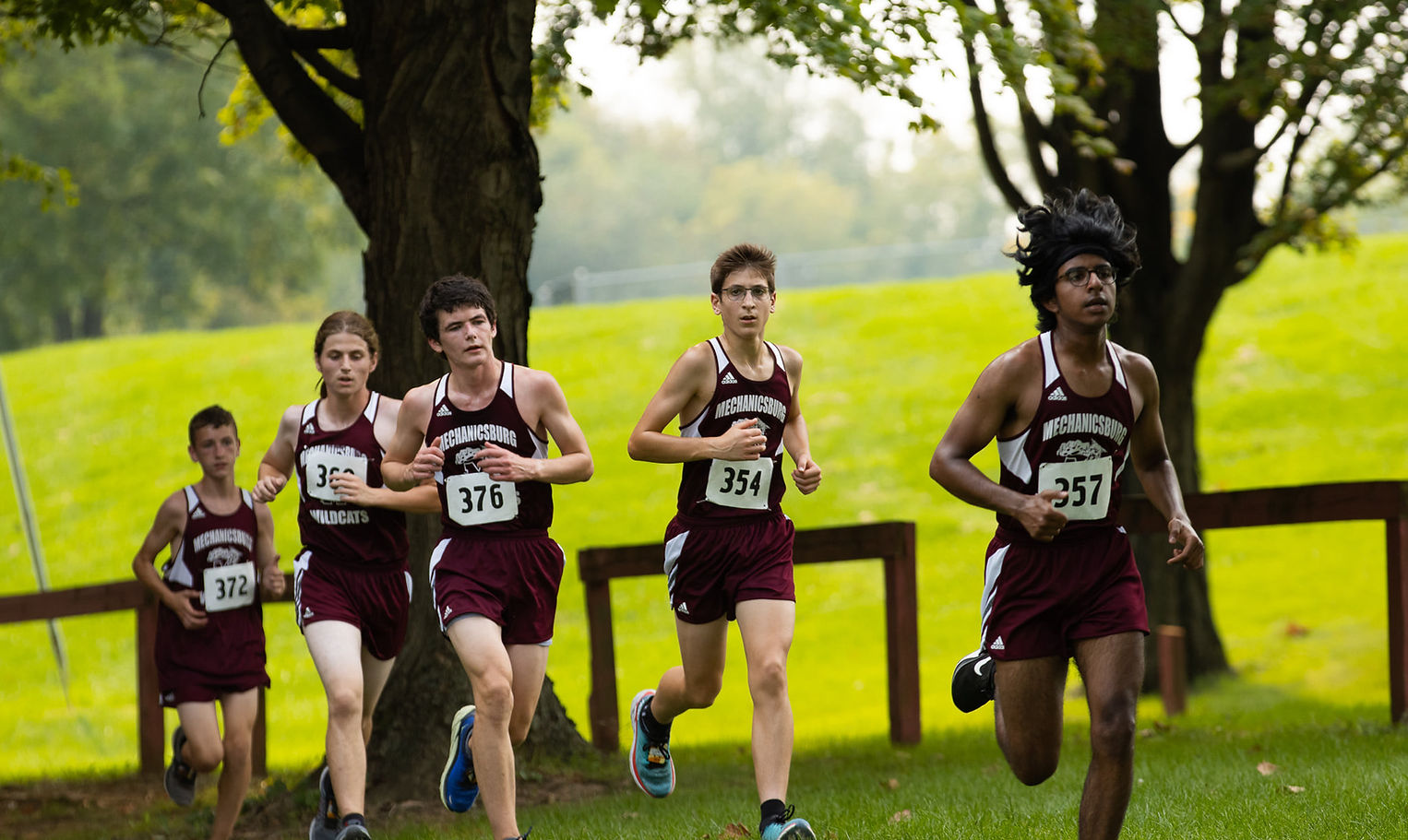 A group of five cross country runners in maroon uniforms are racing through a grassy area with trees in the background.