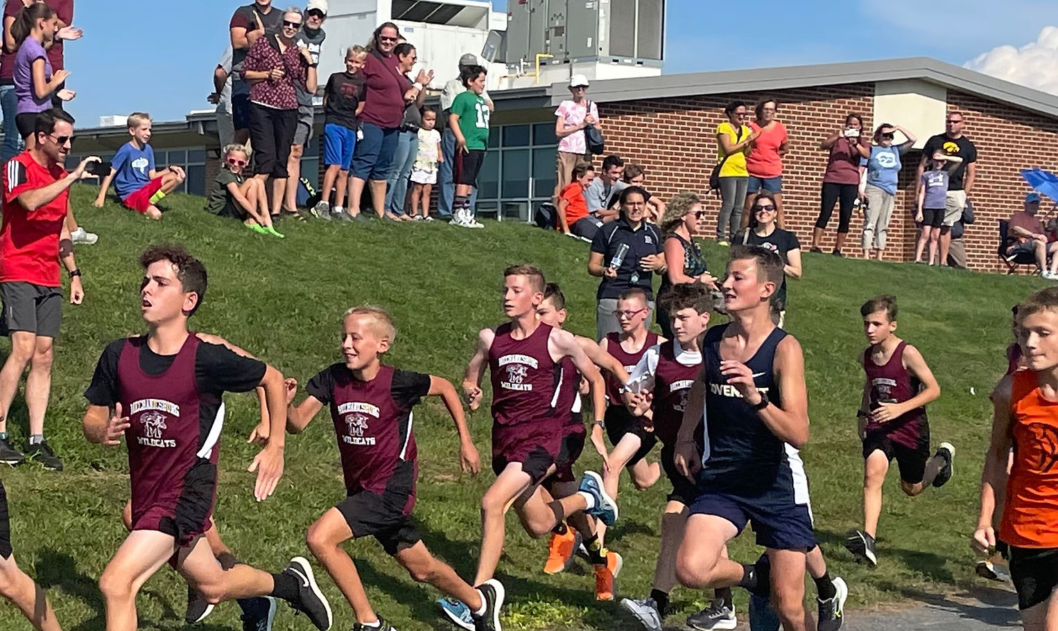 A group of young runners in athletic gear are competing in a race, with spectators cheering from the sidelines on a sunny day.
