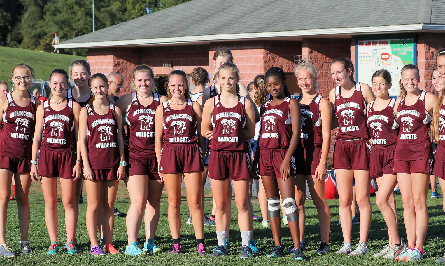 A group of female athletes stands together in matching maroon uniforms, smiling for the camera in a grassy area with a building in the background.