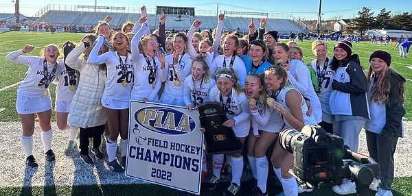 A group of young female athletes celebrating their victory as field hockey champions, holding a trophy and a banner that reads 'PIAA Field Hockey Champions 2022.'