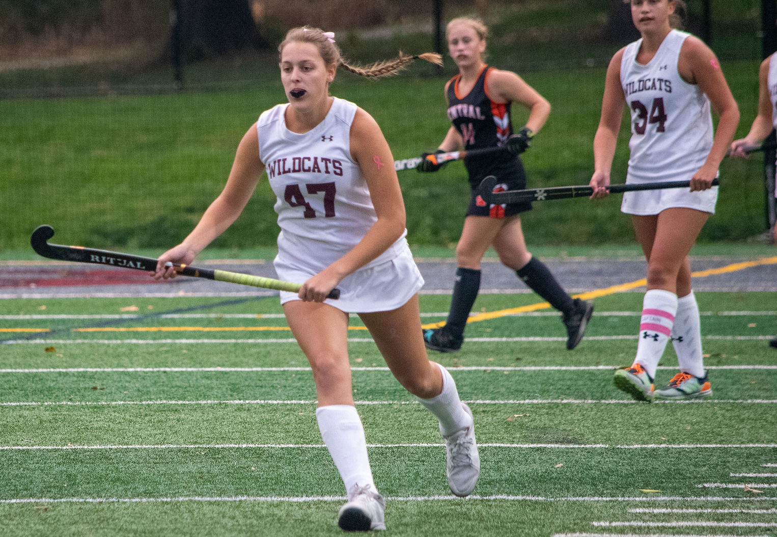 A field hockey player in a white uniform with the number 47 runs with a stick on a turf field, while two other players in the background are positioned for play.
