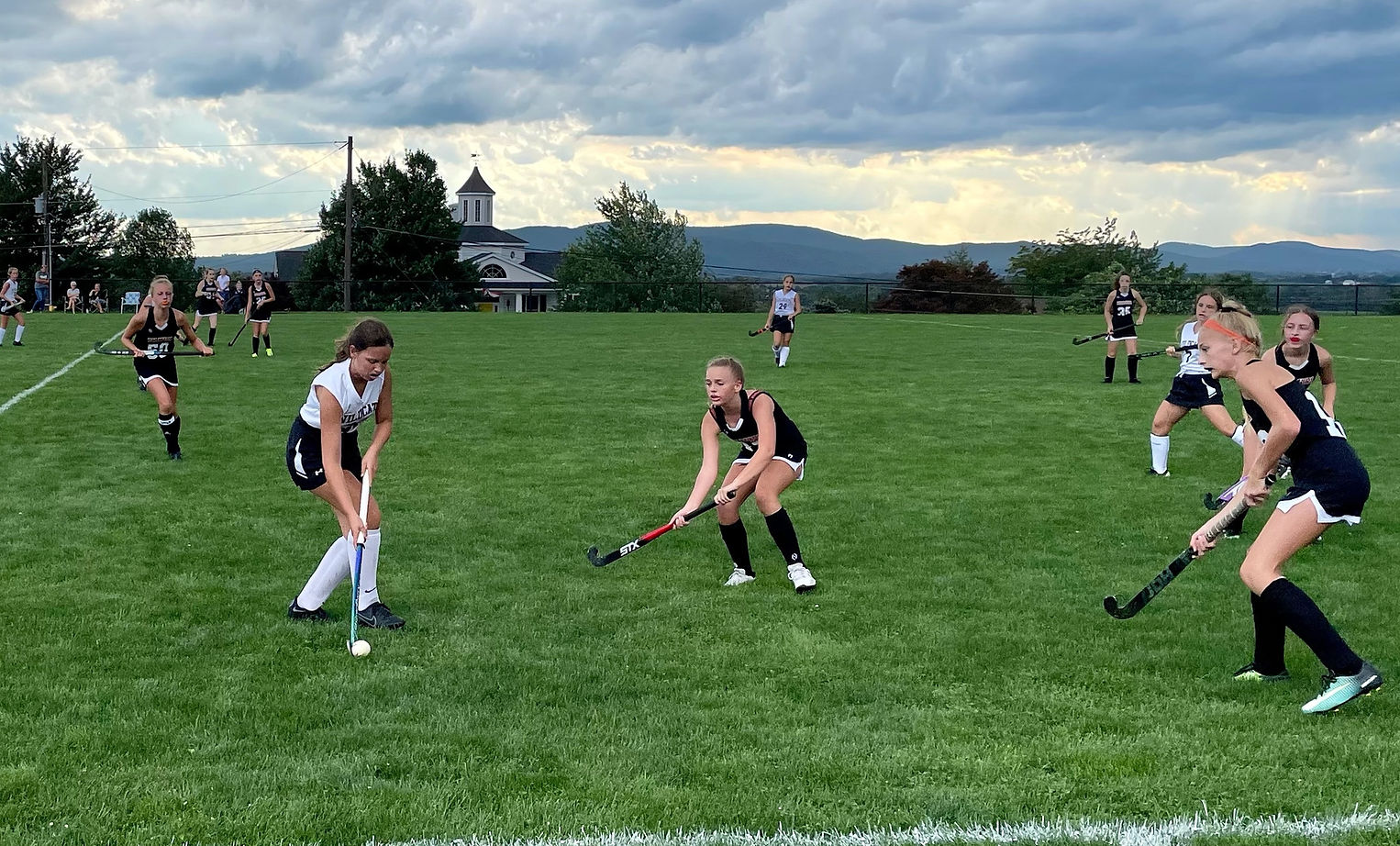 A field hockey match in progress on a grassy field with players in action, set against a backdrop of cloudy skies and distant hills.