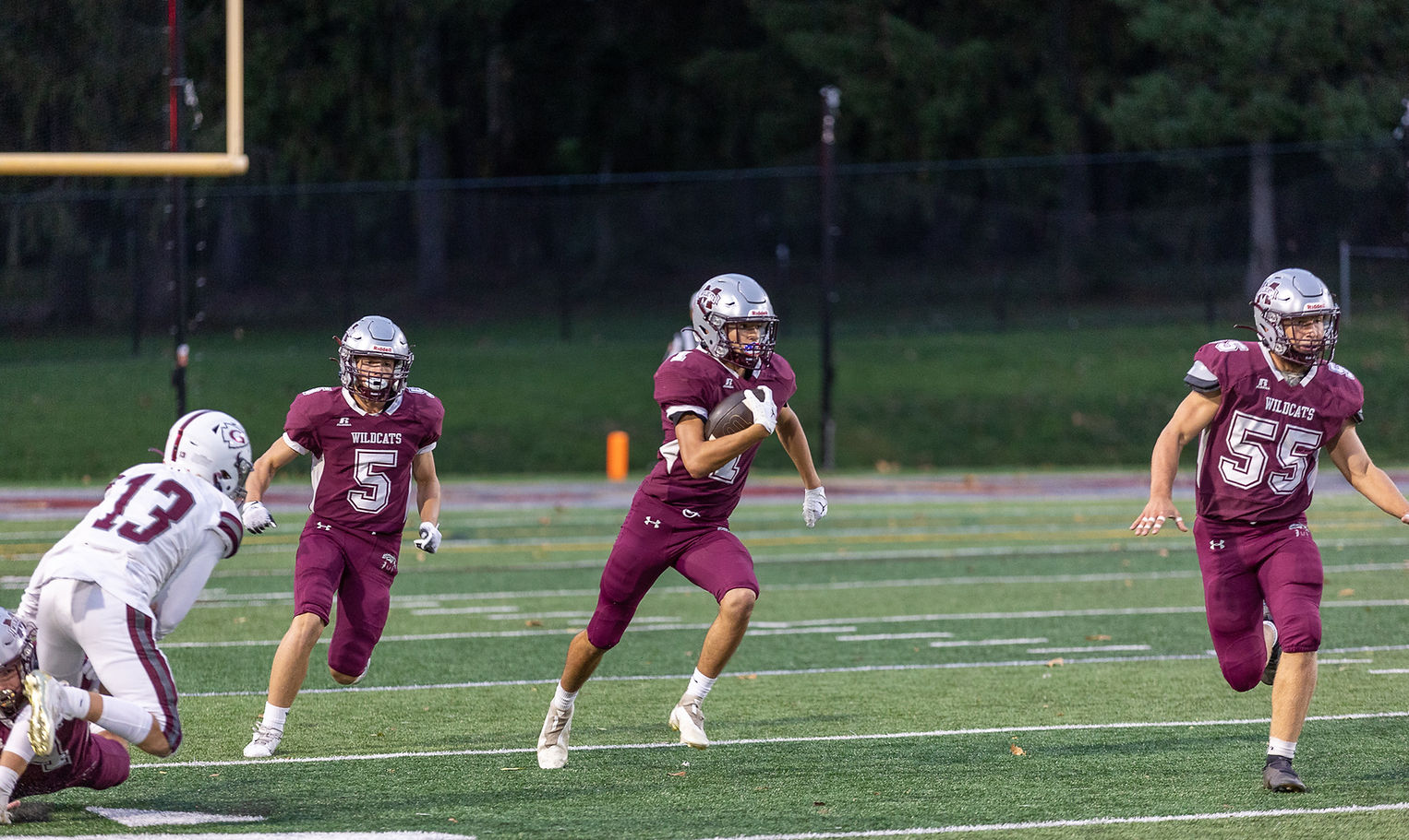 A high school football player in maroon uniform runs with the ball while two teammates support him, and an opposing player attempts to tackle from the ground.