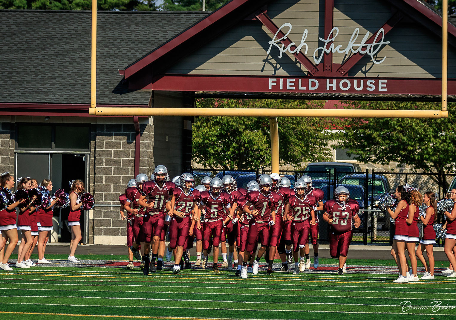 A group of football players in maroon uniforms runs onto the field, flanked by cheerleaders, with a building labeled 'Rich Finkel Field House' in the background.