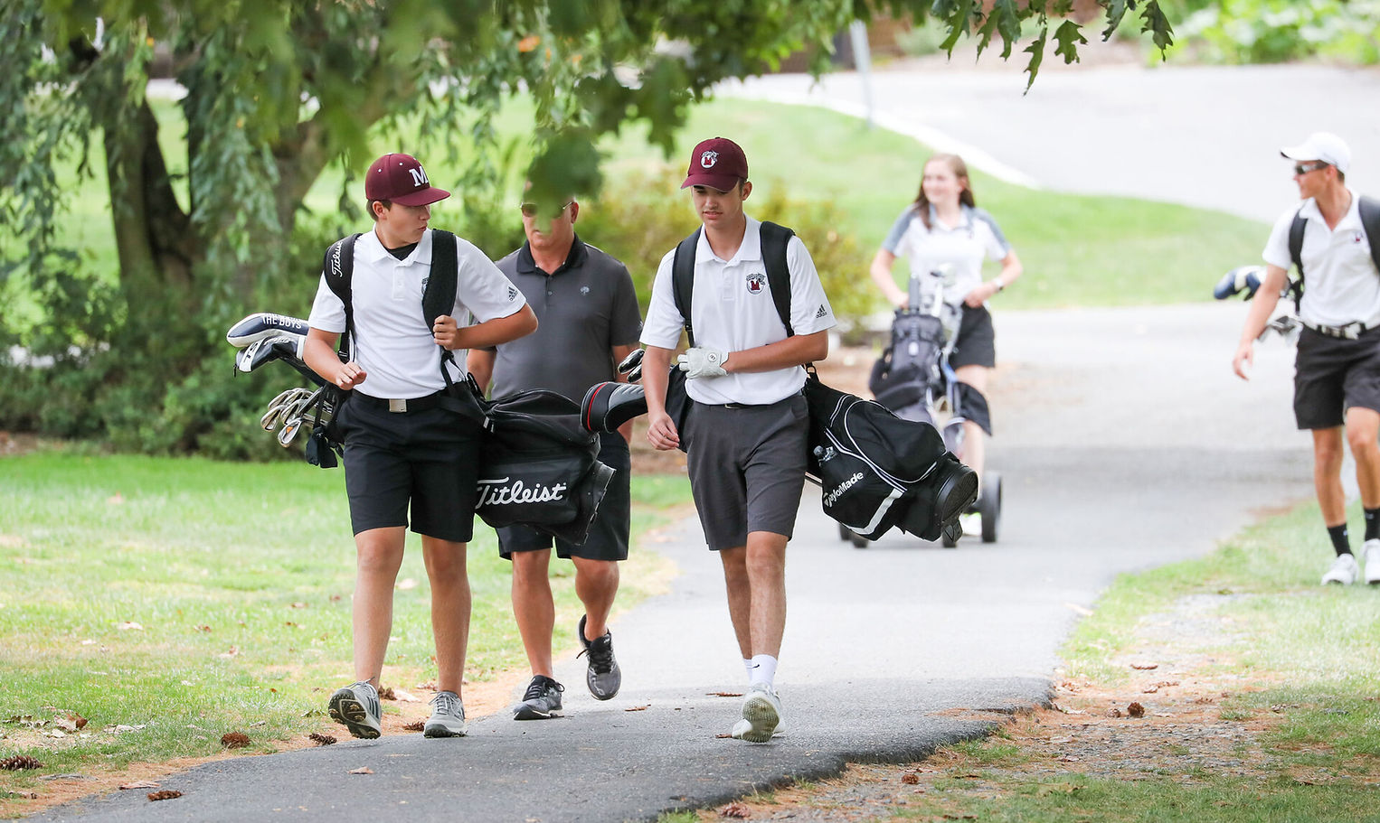 A group of young golfers, dressed in matching white shirts and black shorts, walk along a path carrying golf bags, with trees and a grassy area in the background.