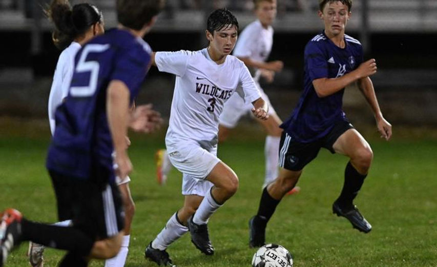 A soccer player in a white uniform dribbles the ball while being pursued by two opponents in dark uniforms during a night game on a grassy field.