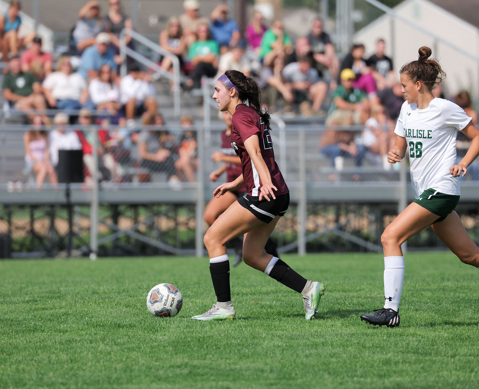 Two female soccer players are competing on a grassy field, with one player in maroon dribbling the ball while the other in white is closely pursuing her, and a crowd is visible in the background.