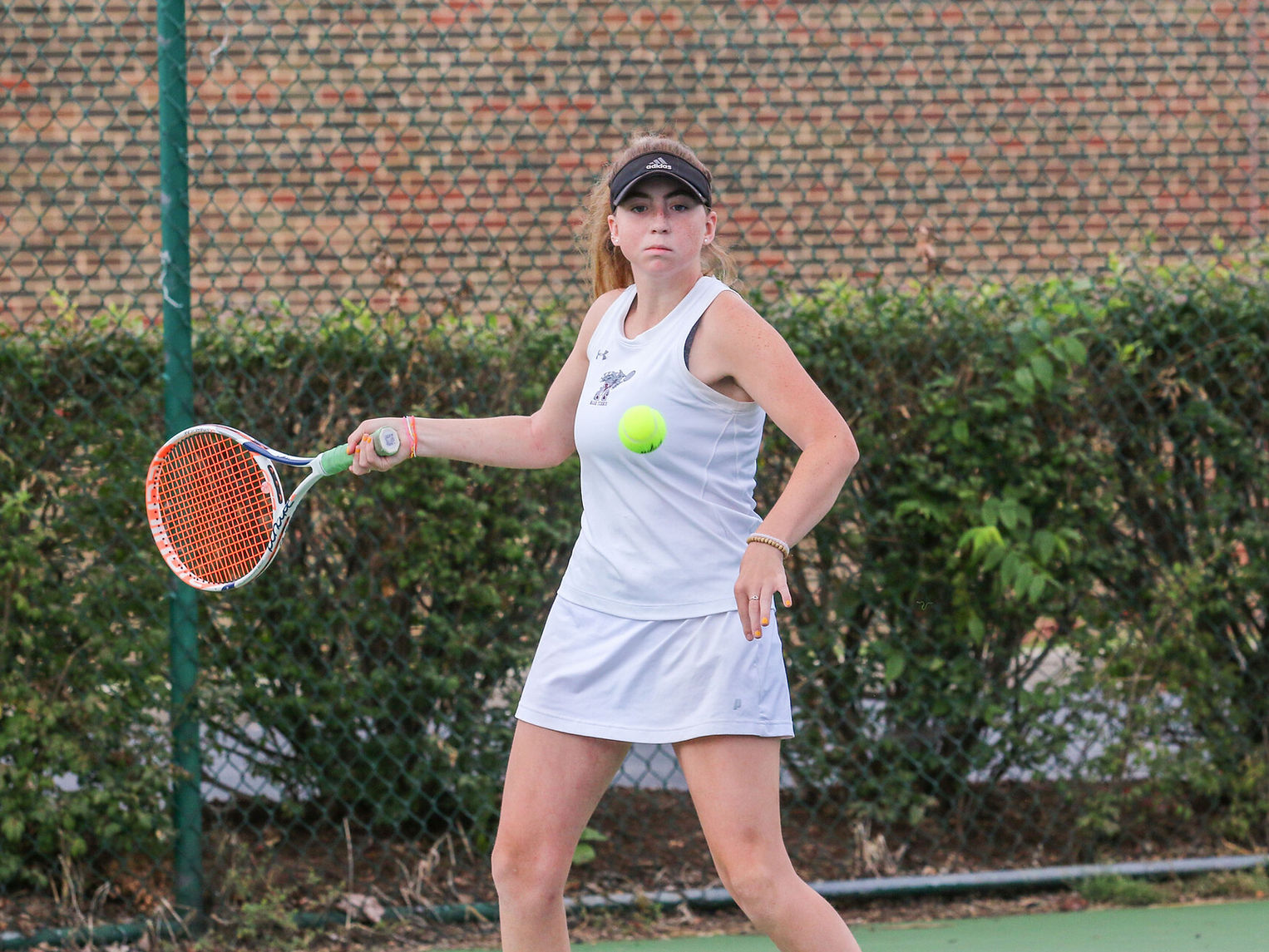 A young female tennis player in a white dress prepares to hit a tennis ball with her racket on a court surrounded by greenery and a brick wall in the background.