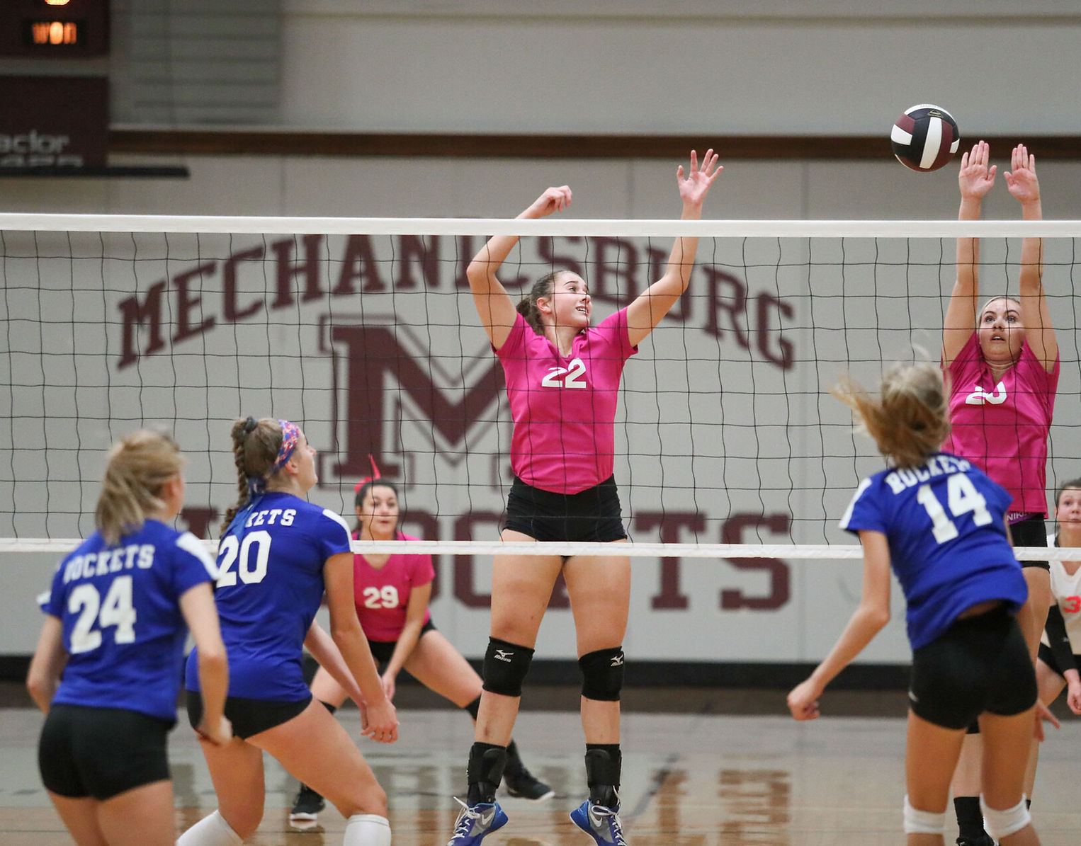 A volleyball player in a pink jersey jumps to spike the ball over the net, while teammates and opponents watch in a gymnasium with a 'Mechanicsburg' logo in the background.