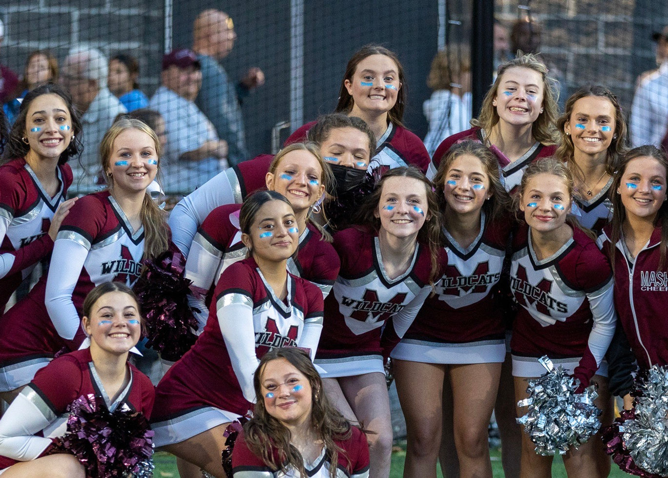 A group of cheerleaders in matching uniforms pose together, smiling and showing enthusiasm, with a crowd visible in the background.