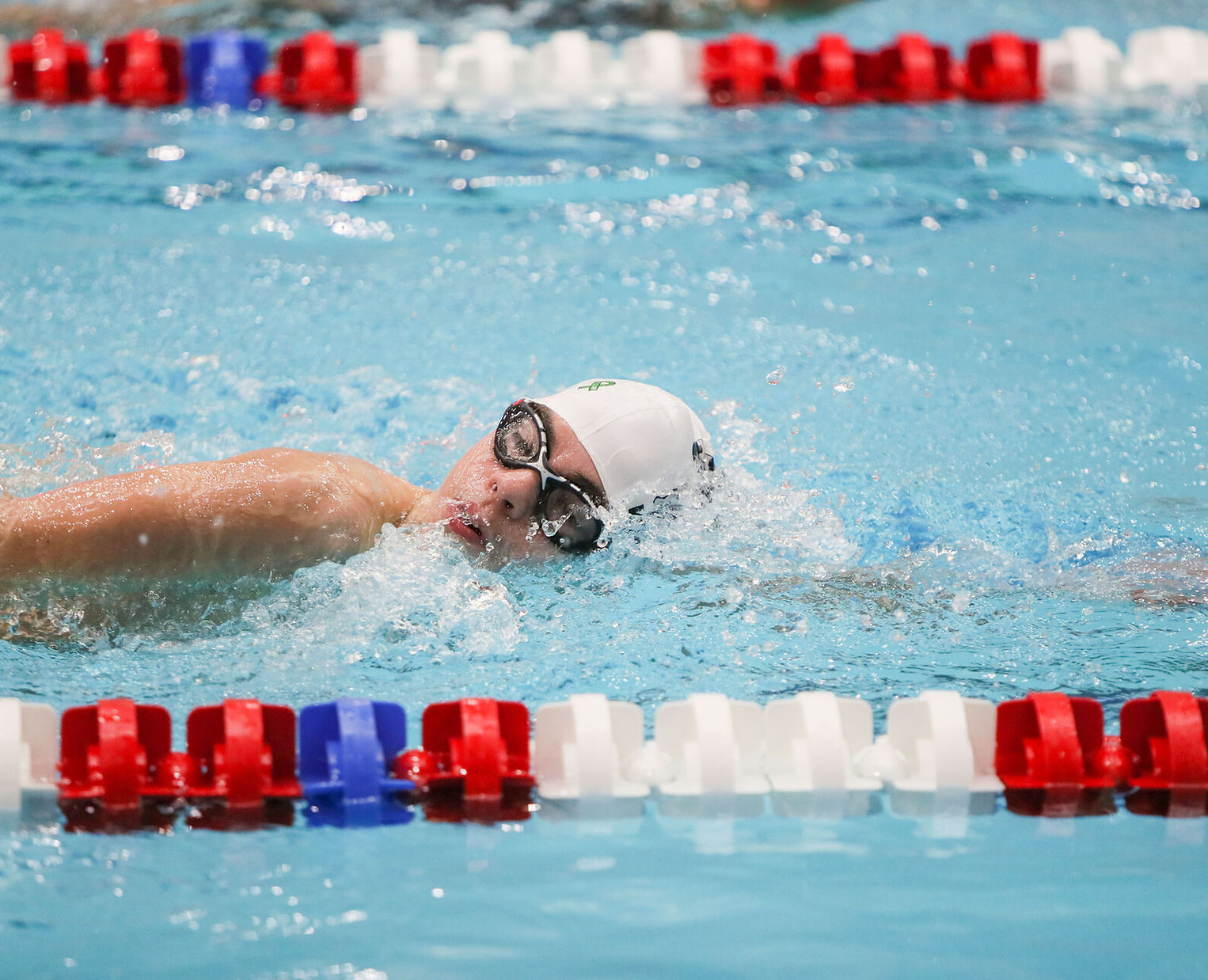 A swimmer in a white cap and goggles is performing a freestyle stroke in a pool, with colorful lane markers visible in the foreground.