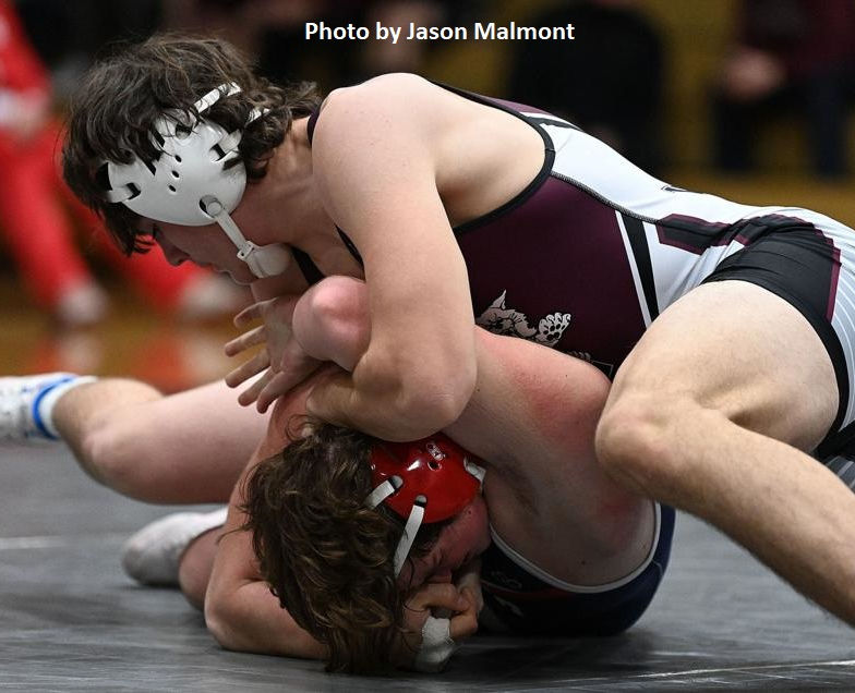 Two high school wrestlers are engaged in a match on a wrestling mat, with one athlete in a white and maroon uniform applying a hold on the other, who is wearing a red uniform.