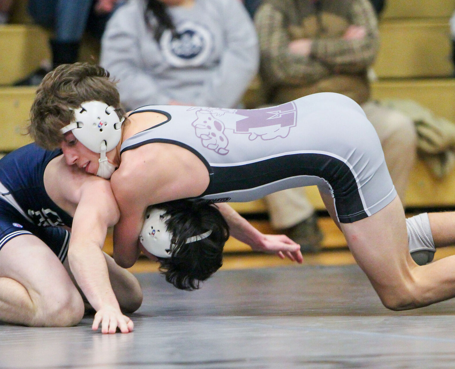 Two high school wrestlers are engaged in a match on a wrestling mat, with one athlete in a gray uniform attempting to gain control over the other, who is in a blue uniform.