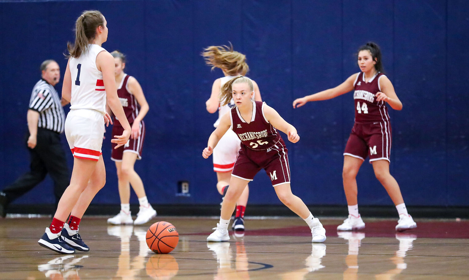 A basketball game in progress featuring female players in maroon and white uniforms, with one player dribbling the ball while others are positioned defensively.