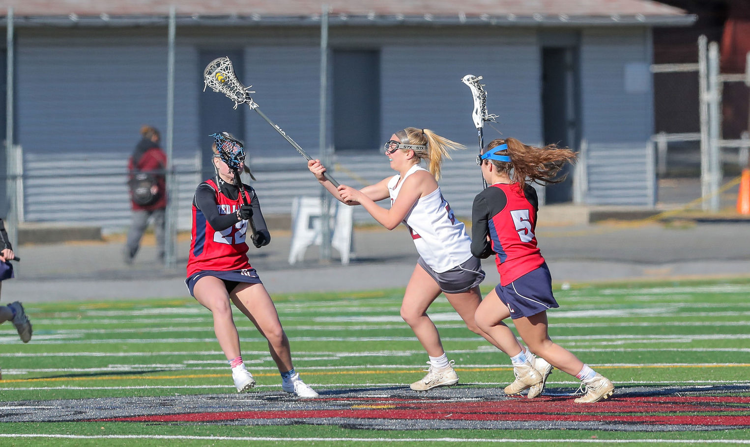 A female lacrosse player in a white uniform is attempting to pass the ball while being closely guarded by two opponents in red uniforms on a grassy field.