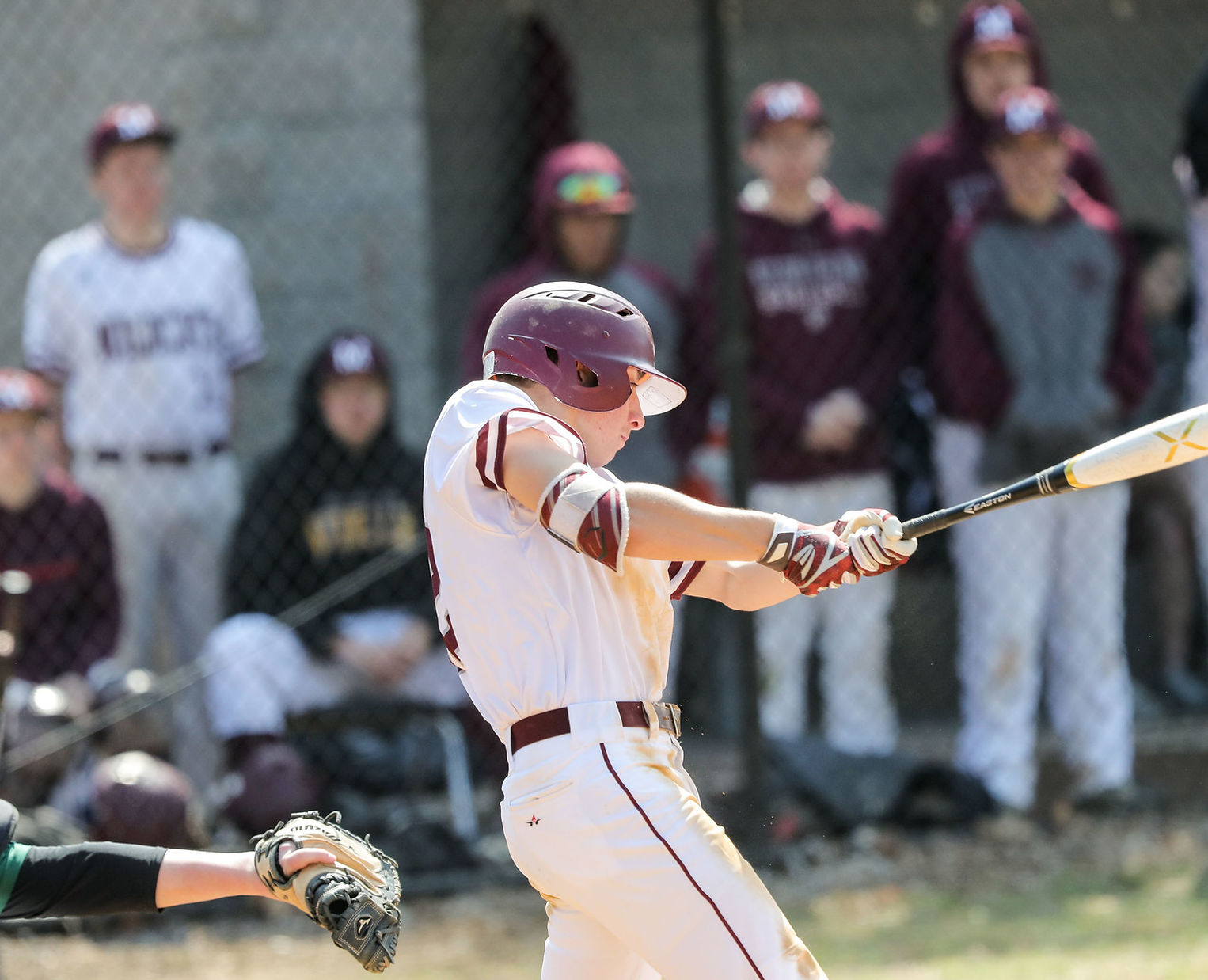 A baseball player in a white uniform swings a bat during a game, with teammates in the background watching from the dugout.