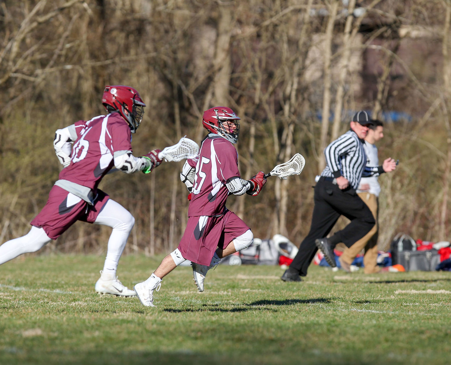 Two lacrosse players in maroon uniforms sprint across a grassy field while a referee runs in the opposite direction, with trees in the background.