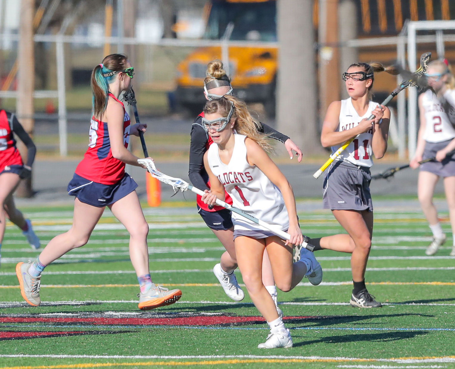 A group of female lacrosse players are engaged in a game on a field, with one player in the foreground running with the ball while others are in motion behind her.