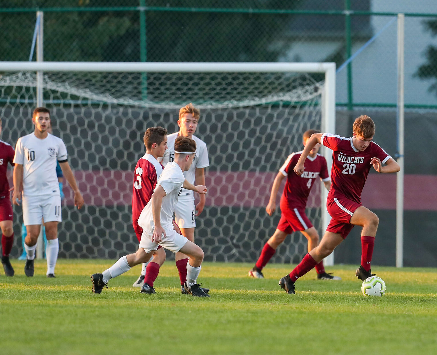 A soccer match in progress, featuring players in red and white uniforms competing for the ball on a grassy field with a goal in the background.