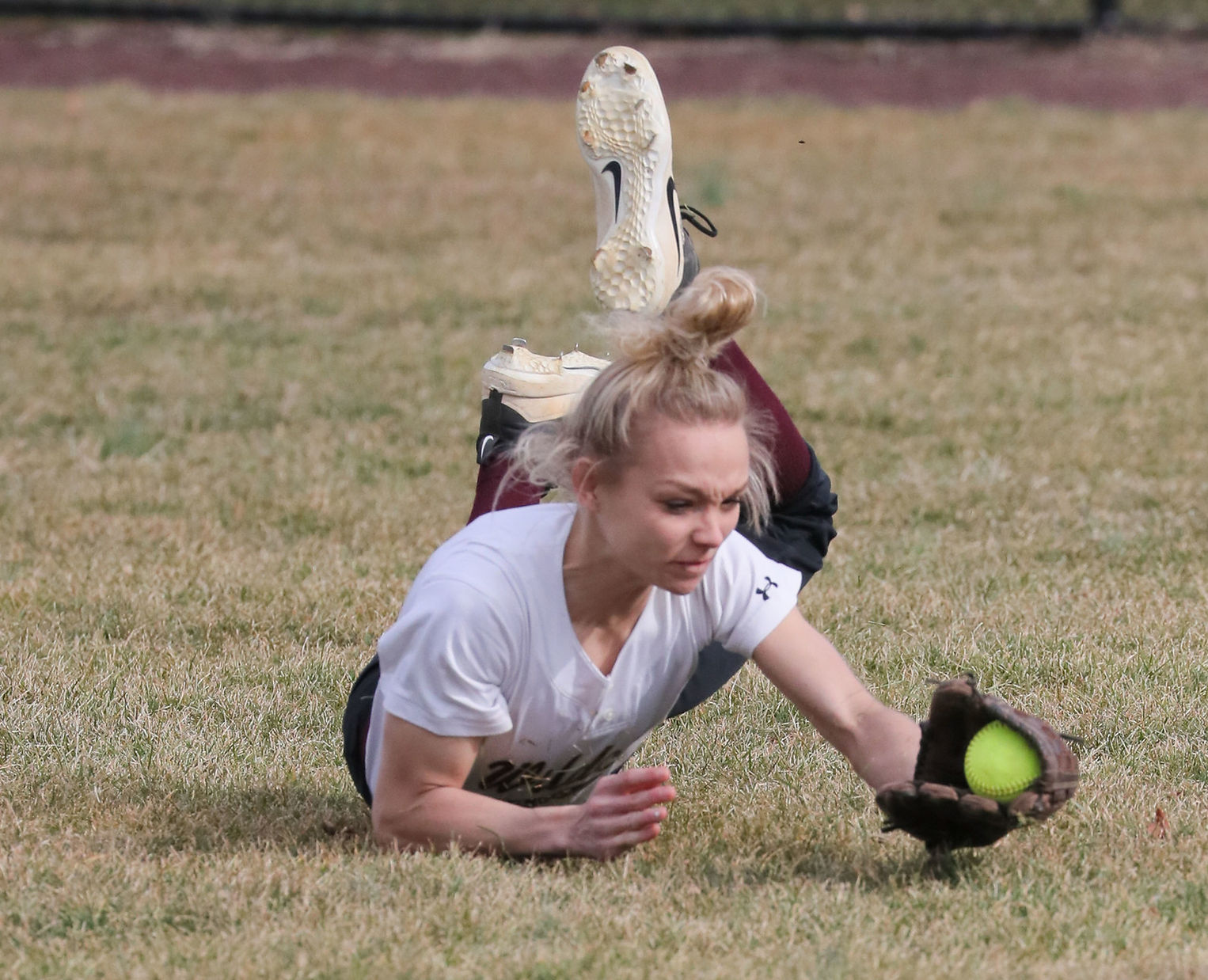 A young athlete dives on the grass to catch a softball, with her glove extended and a focused expression on her face.