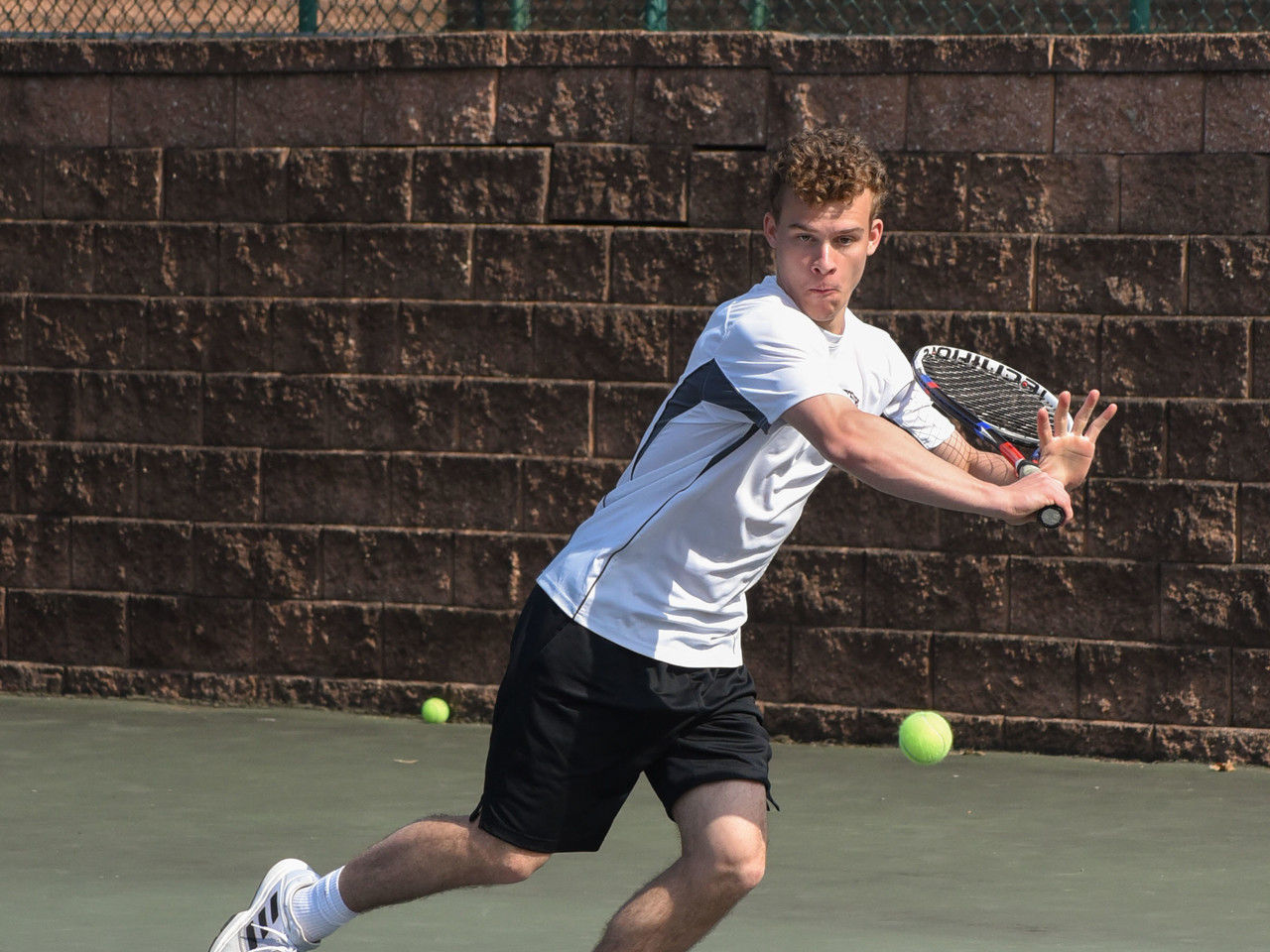 A young male tennis player is in mid-swing on a court, with tennis balls scattered around him and a textured wall in the background.