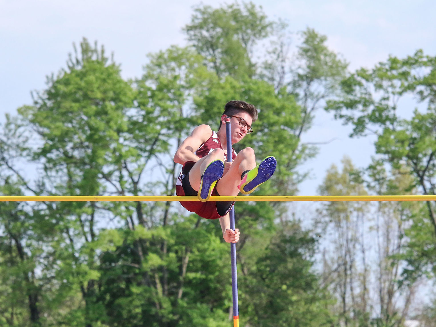 A pole vaulter in mid-air, gripping a pole, as they clear a bar against a backdrop of trees and blue sky.