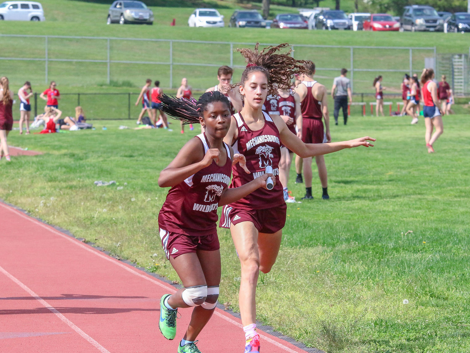 Two female track athletes in maroon uniforms are competing in a relay race, with one runner passing the baton to the other on a running track, while spectators and other athletes are visible in the background.