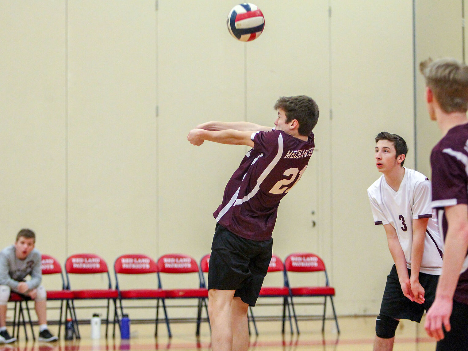A volleyball player in a maroon jersey prepares to receive a serve, while teammates watch from the sidelines in a gymnasium setting.