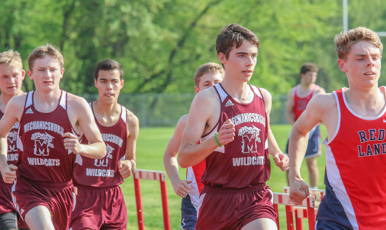 A group of high school male runners in athletic uniforms compete in a track event, with one runner in the foreground leading the pack.