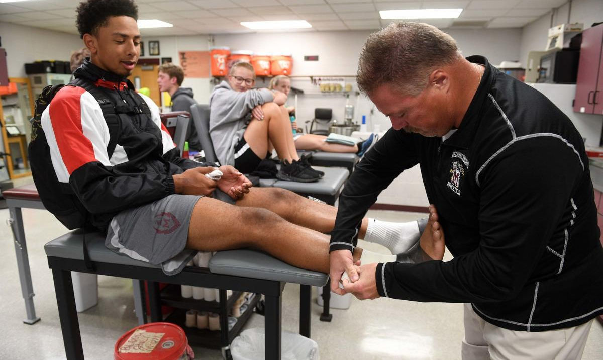 A sports trainer is applying treatment to an athlete's ankle while another athlete sits nearby, both in a training room setting.