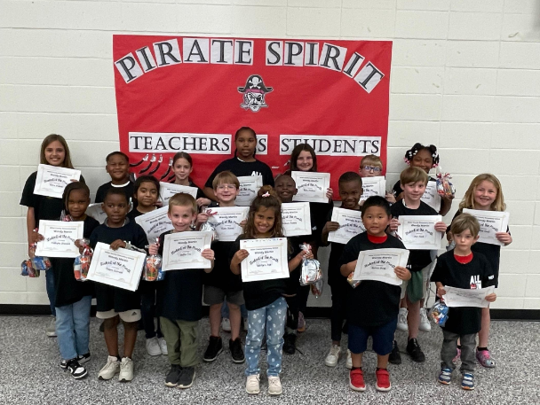A group of students holding certificates stands in front of a red banner that reads 'Pirate Spirit' and 'Teachers & Students.'