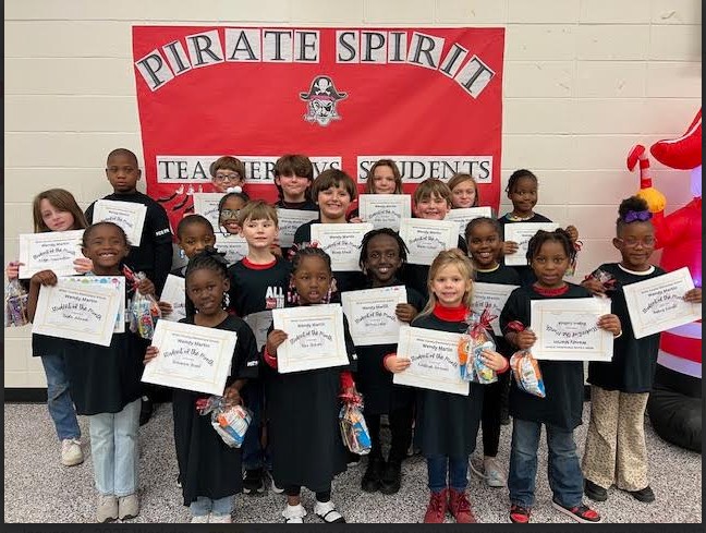 A group of children holding certificates stands in front of a red banner that reads 'Pirate Spirit' in a school setting.
