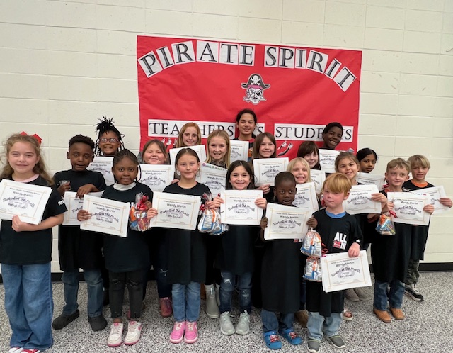 A group of children holding certificates and small bags, standing in front of a red banner that reads 'Pirate Spirit' in a school setting.