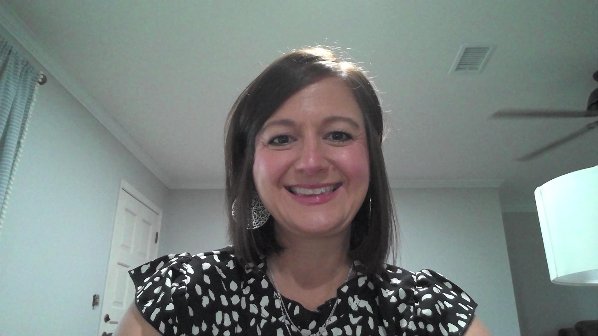 A woman with shoulder-length brown hair smiles at the camera while wearing a black and white patterned blouse, with a light-colored wall and a ceiling fan visible in the background.