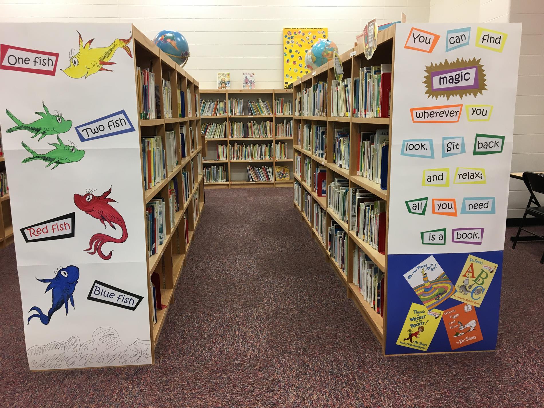 A view of a library aisle with wooden bookshelves filled with books, decorated with colorful fish illustrations and motivational phrases on the ends of the shelves.