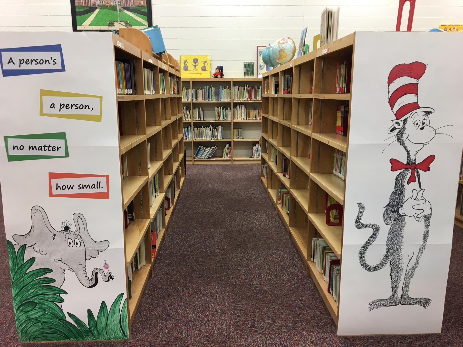 A view of a library aisle with wooden bookshelves on either side, featuring colorful illustrations of a cat in a hat and an elephant, along with a quote about the importance of every person.