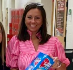 A woman wearing a pink and white striped shirt smiles while holding a box of snacks in a school hallway.