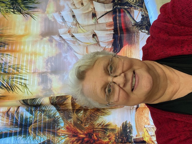 A woman with short gray hair and glasses smiles in front of a colorful backdrop featuring palm trees and a sailing ship.