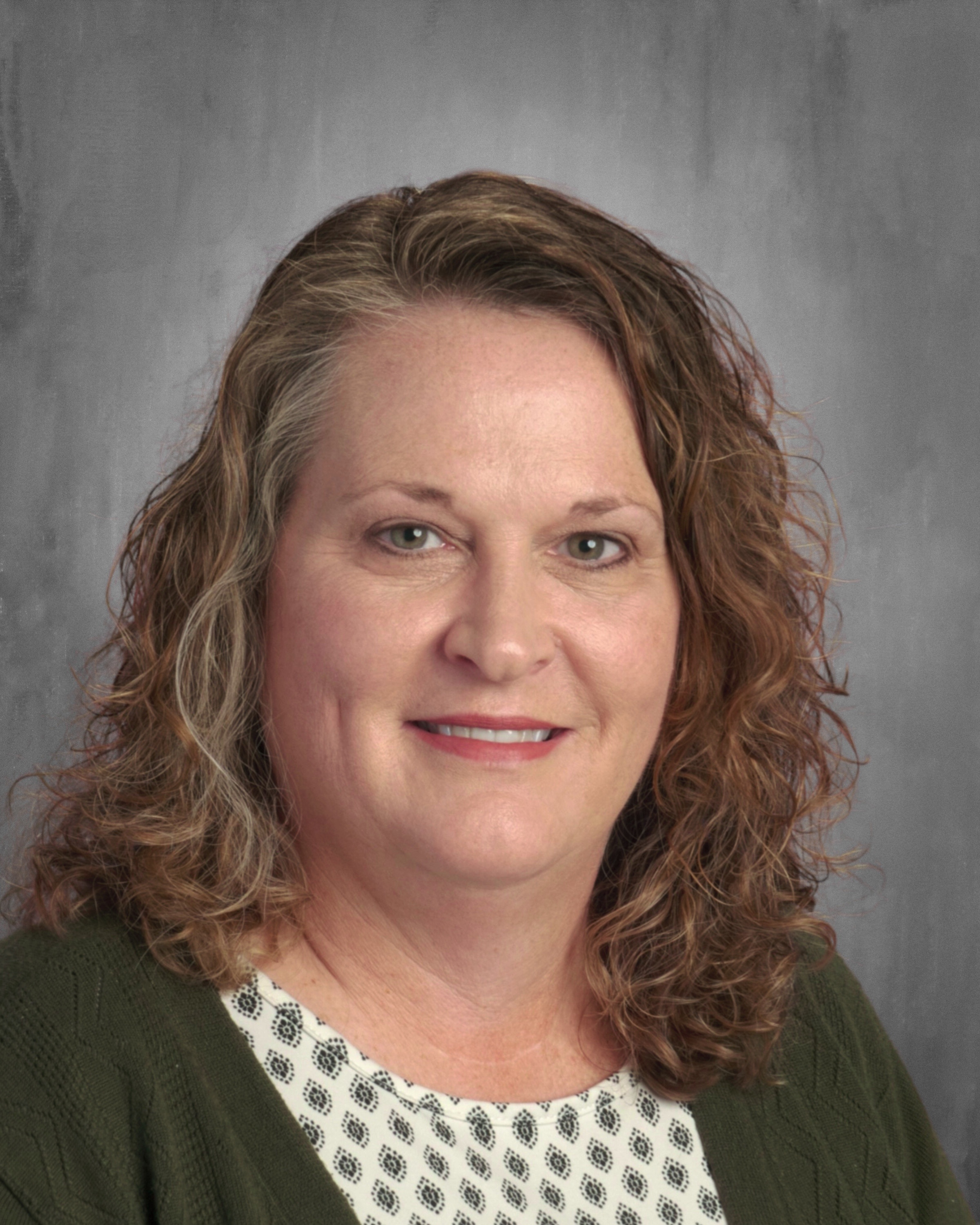 A woman with curly hair, wearing a green cardigan over a patterned blouse, smiles for the camera against a gray background.