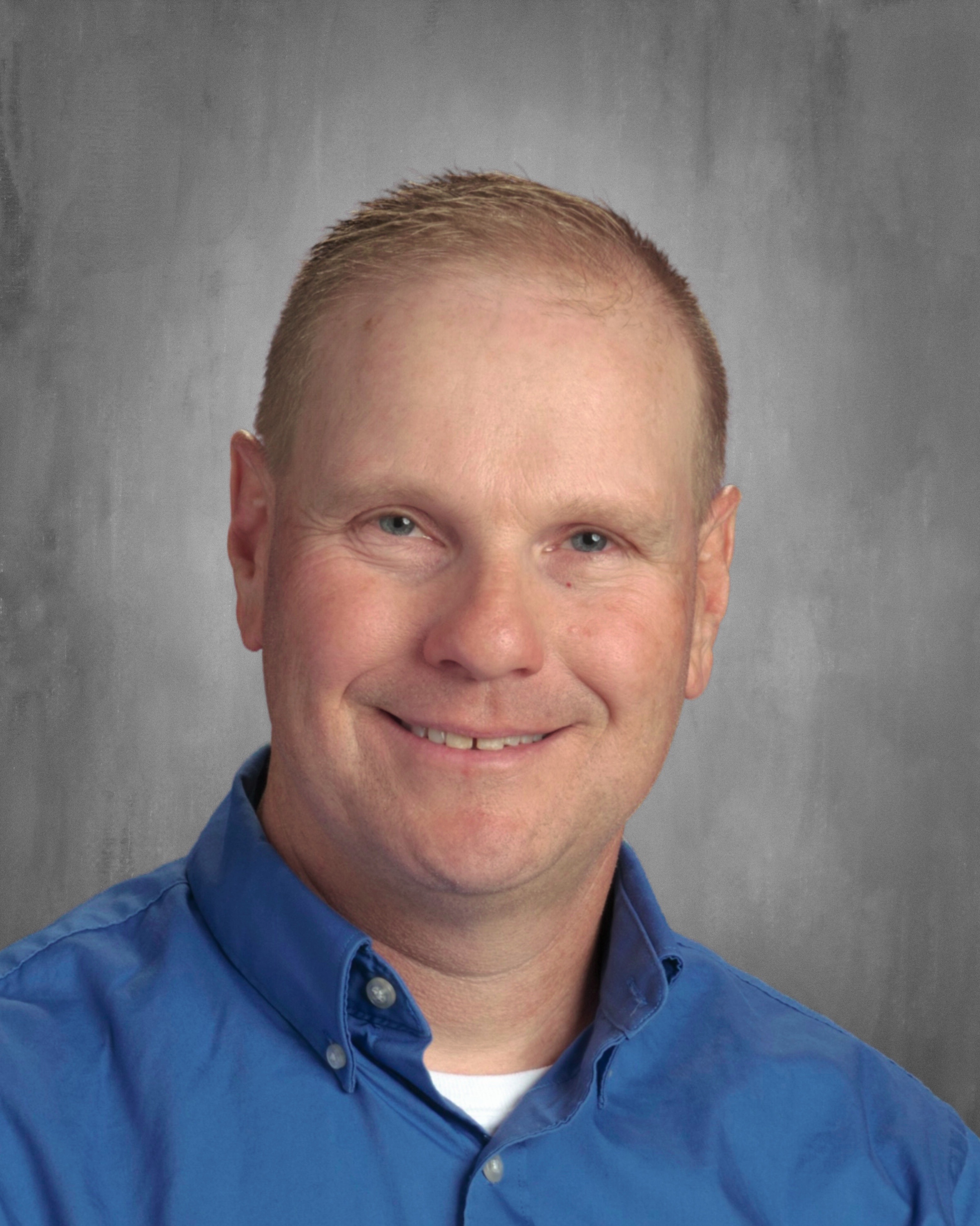A smiling man with short hair wearing a blue collared shirt against a gray background.