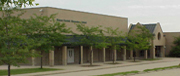A view of a brick building with a flat roof, surrounded by small trees and a paved pathway, likely a school or community center.